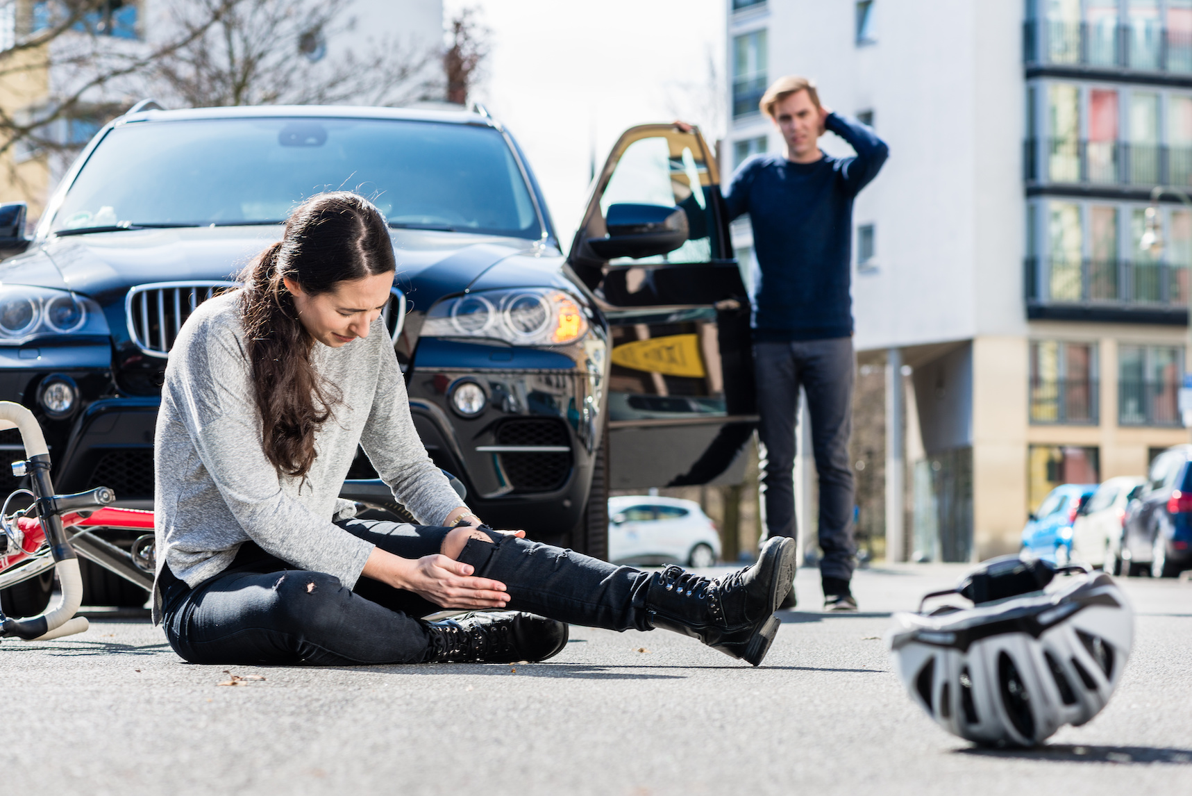 Full length of a young female bicyclist fallen down on street with serious injuries after traffic accident with the 4x4 car of a young man, who looks concerned.Depicts fault in car accidents.